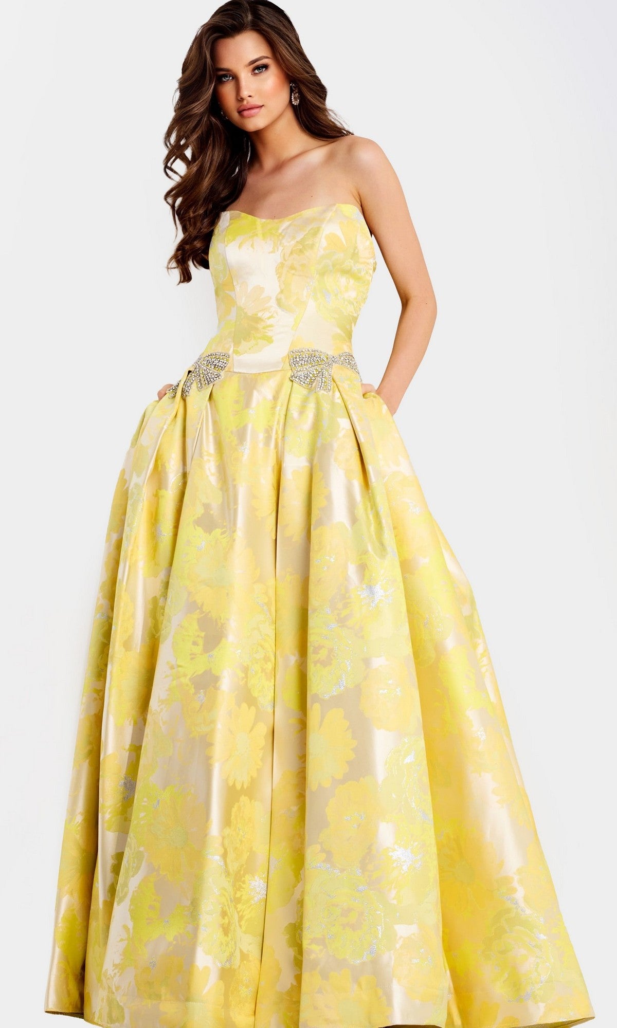 A woman in the Jovani 49502 Strapless Long Yellow Prom Ball Gown with silver waist embellishments stands against a light background.